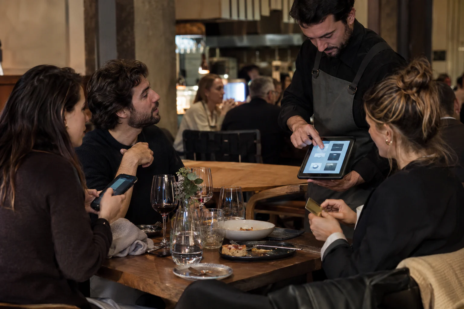 Camarero mostrando una tableta a un grupo de comensales mientras una clienta sostiene su tarjeta